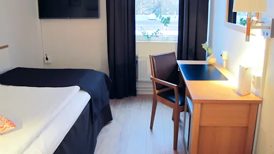 Et pent hotellrom med en seng til venstre, en veggmontert TV rett overfor og et skrivebord i tre med stol nær et vindu med mørke gardiner.