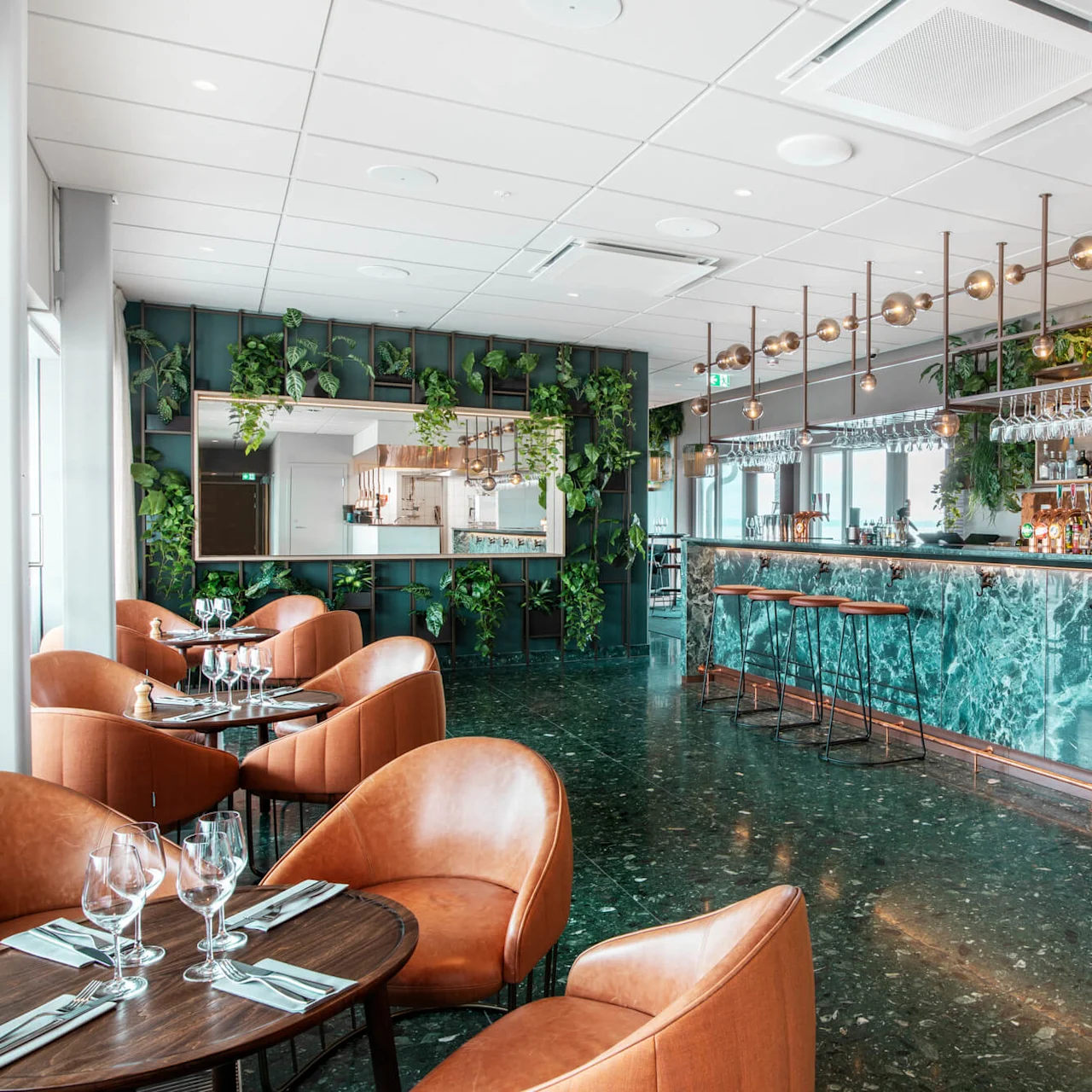 Elegant bar med blå marmordisk, omgitt av brune lærstoler og bord, med hengende glass. Frodig grønne planter dekorerer veggene og skaper en moderne, sofistikert stemning.