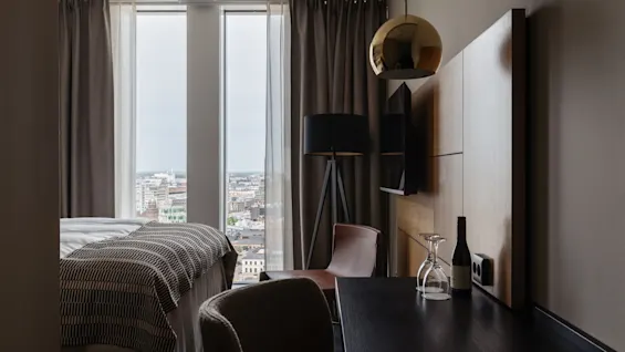 Et moderne hotellrom med en komfortabel seng, et skrivebord med en flaske vin og et stort vindu med utsikt over en by.