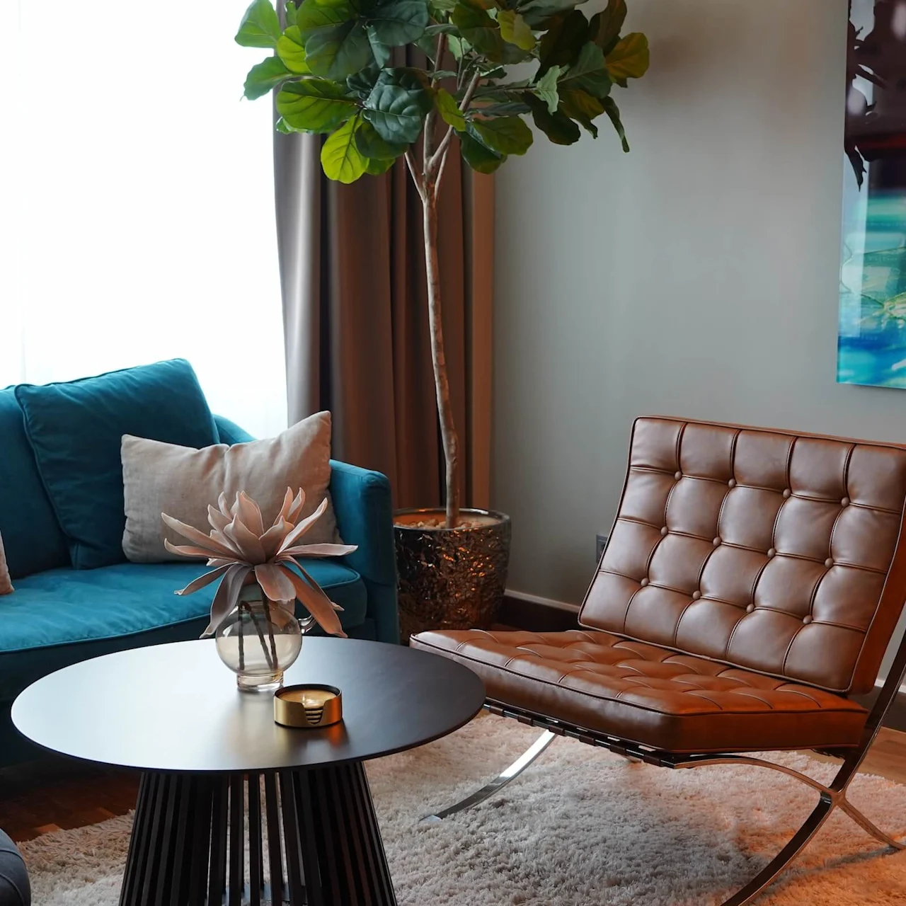 A blue sofa holds pillows beside a black round table with a vase, situated near a brown chair and a tall plant, under soft indoor lighting.