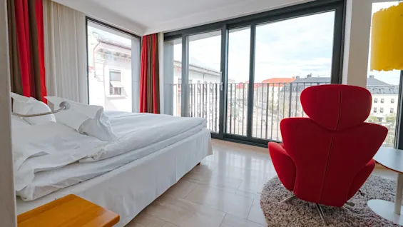 A red armchair faces large floor-to-ceiling windows with city views in a bright room. A neatly made bed and colorful curtains enhance the modern aesthetic.