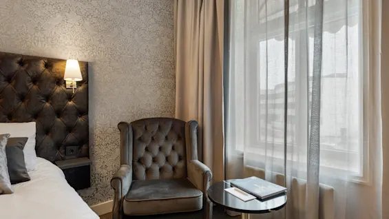 Et hyggeligt hotelværelse med en komfortabel seng, en lænestol og et lille bord ved et vindue med tynde gardiner.