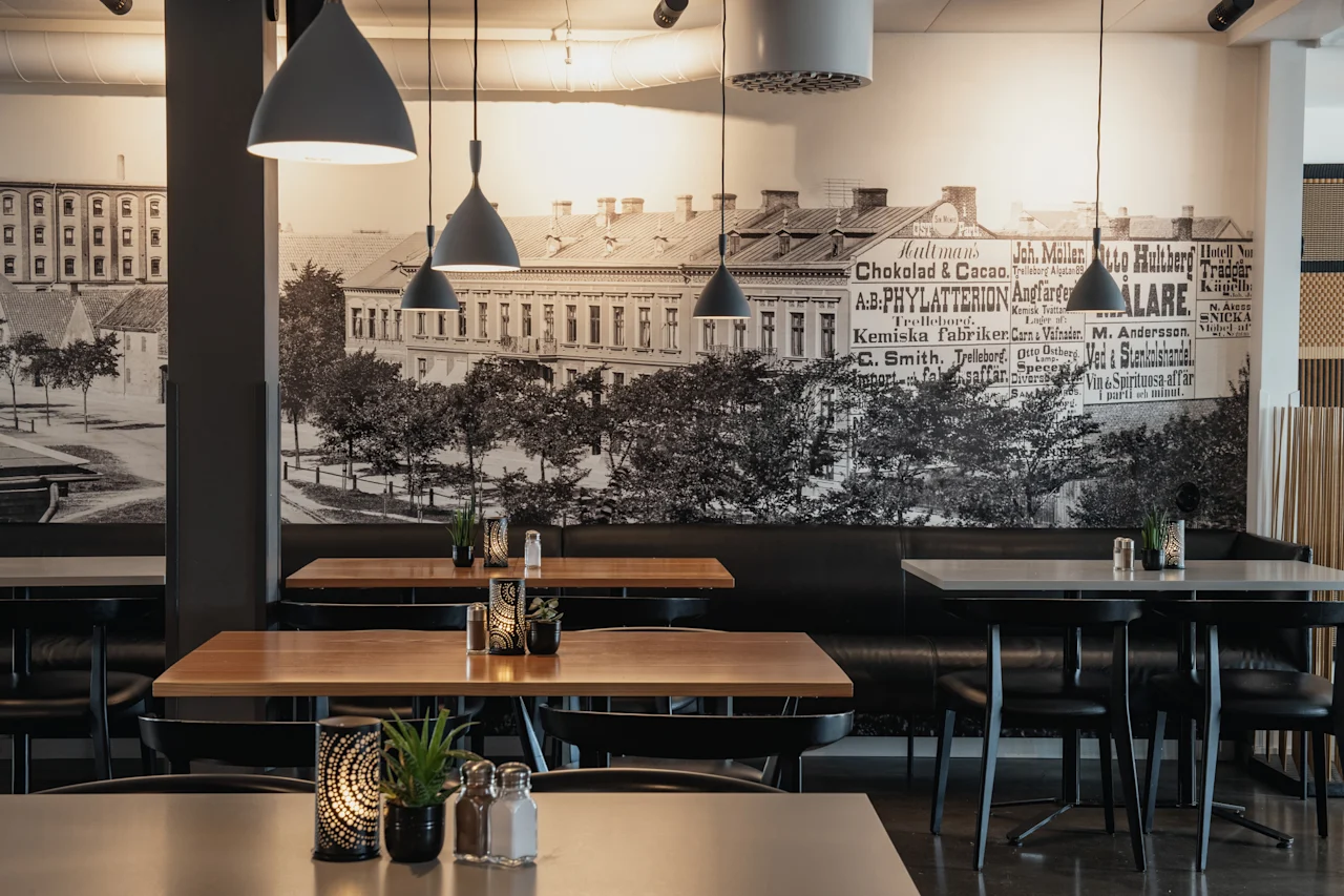 Moderni ravintola puupöydillä, mustilla tuoleilla ja riippuvalaisimilla. Suuri seinämaalaus vanhasta kaupungista vintage-mainoksilla.