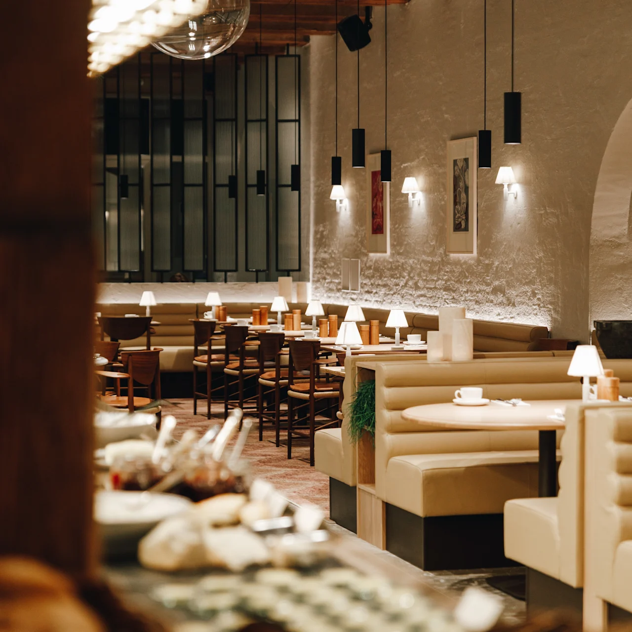 Restaurant interior features cozy beige booths and wooden tables, set under warm hanging lights in a spacious room with brick walls and art decor.