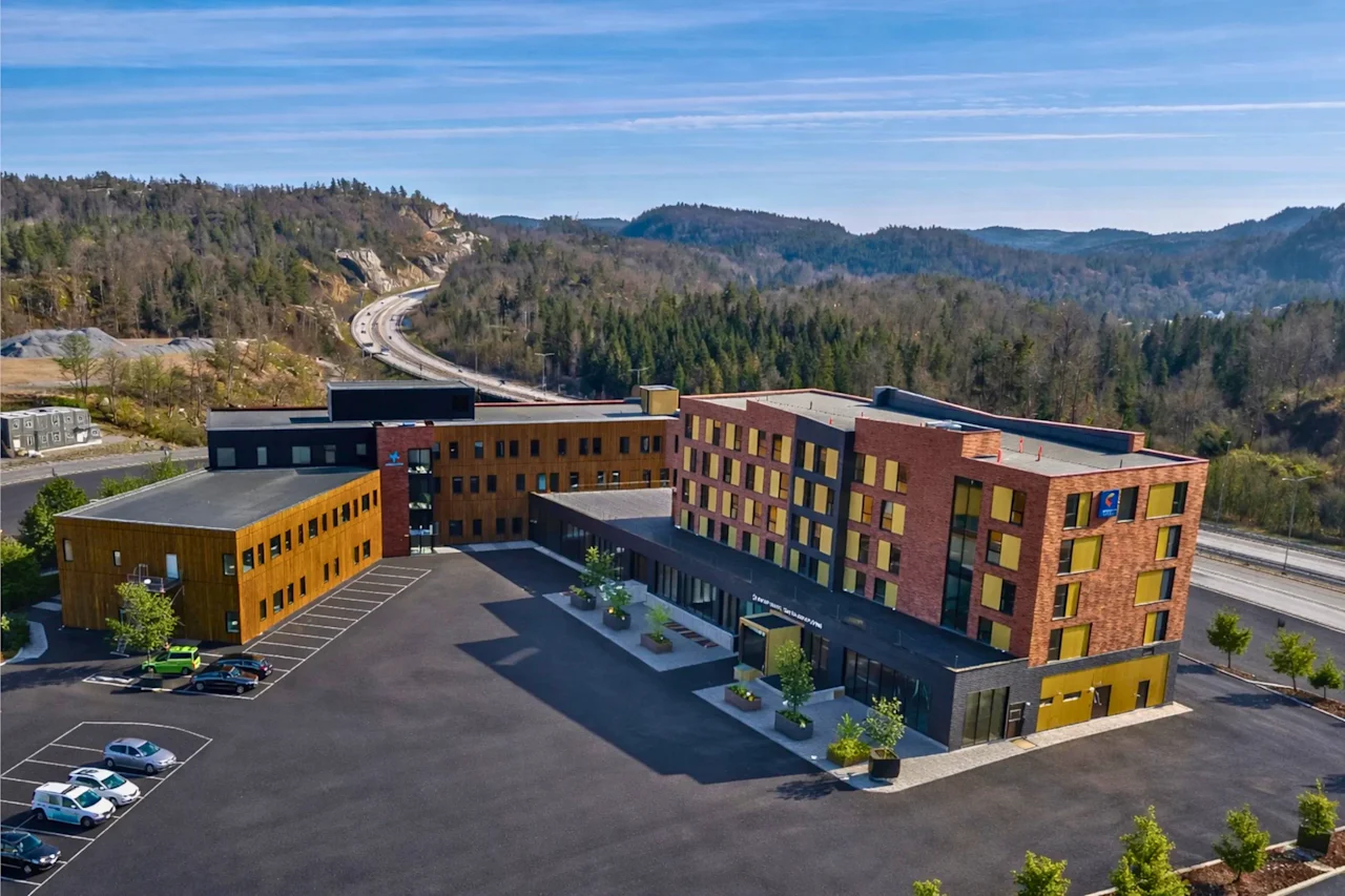 En moderne hotellbygning med en stor parkeringsplass, mot et bakteppe av skogkledde åser og en svingete vei.
