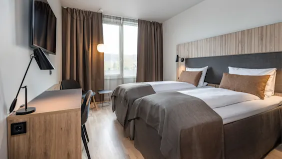 Et værelse med to pænt opredte enkeltsenge, brune nuancer, et skrivebord af træ med lampe og vægmonteret TV; et vindue med trukne gardiner giver naturligt lys.