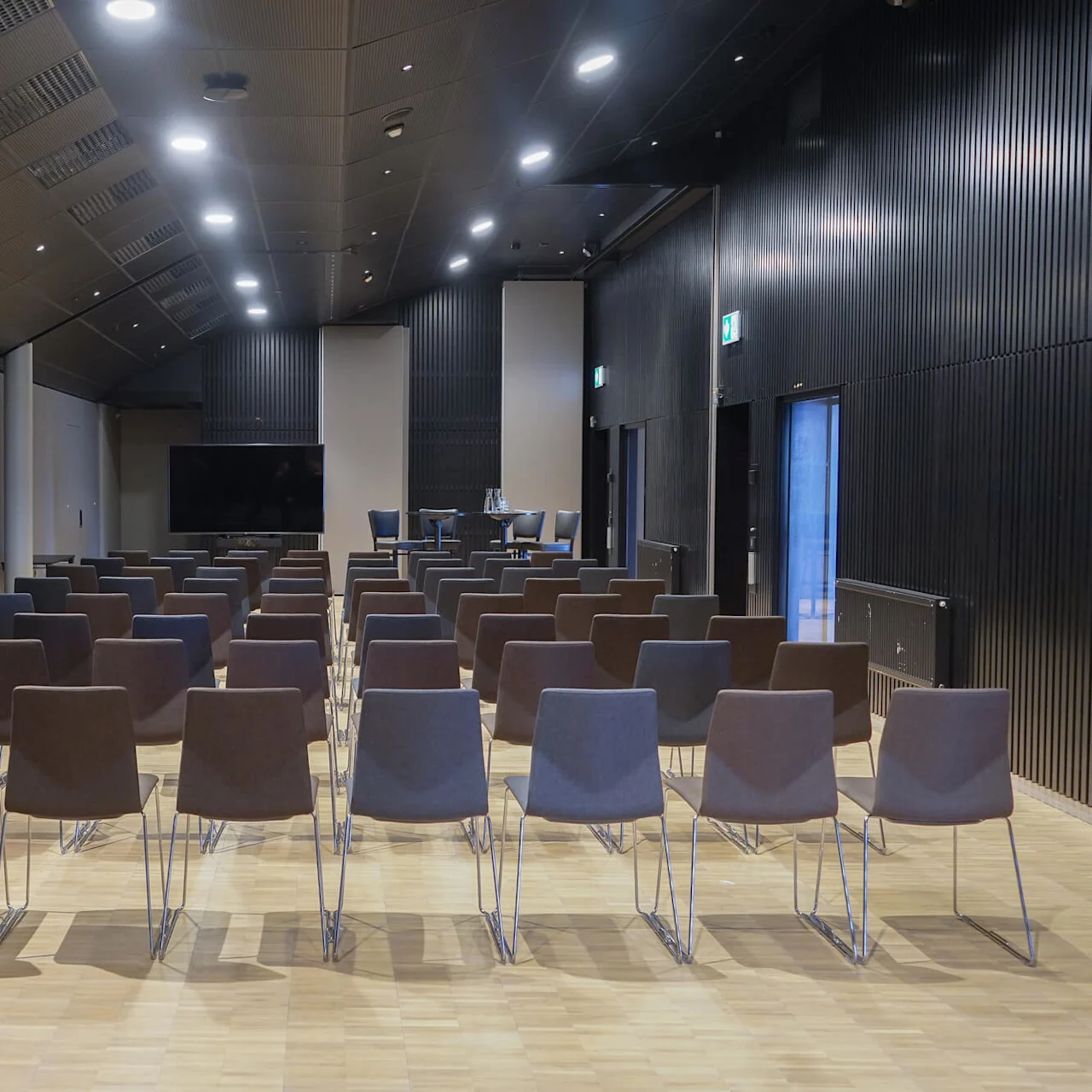Rader med tomme stoler peker mot et presentasjonsområde med et bord og skjerm i et moderne, godt opplyst konferanserom med tregulv og teksturerte vegger.