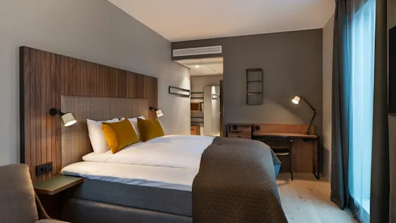 En pent oppredd seng med gule puter står mot en trepanel i et moderne hotellrom. En skrivebordslampe lyser opp en arbeidsbord. Store vinduer med gardiner slipper inn naturlig lys.