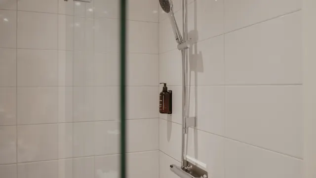 Duschhuvud fäst vid kaklad vägg, ovanför en liten brun flaska; omgiven av en klar glasruta i ett modernt badrum.