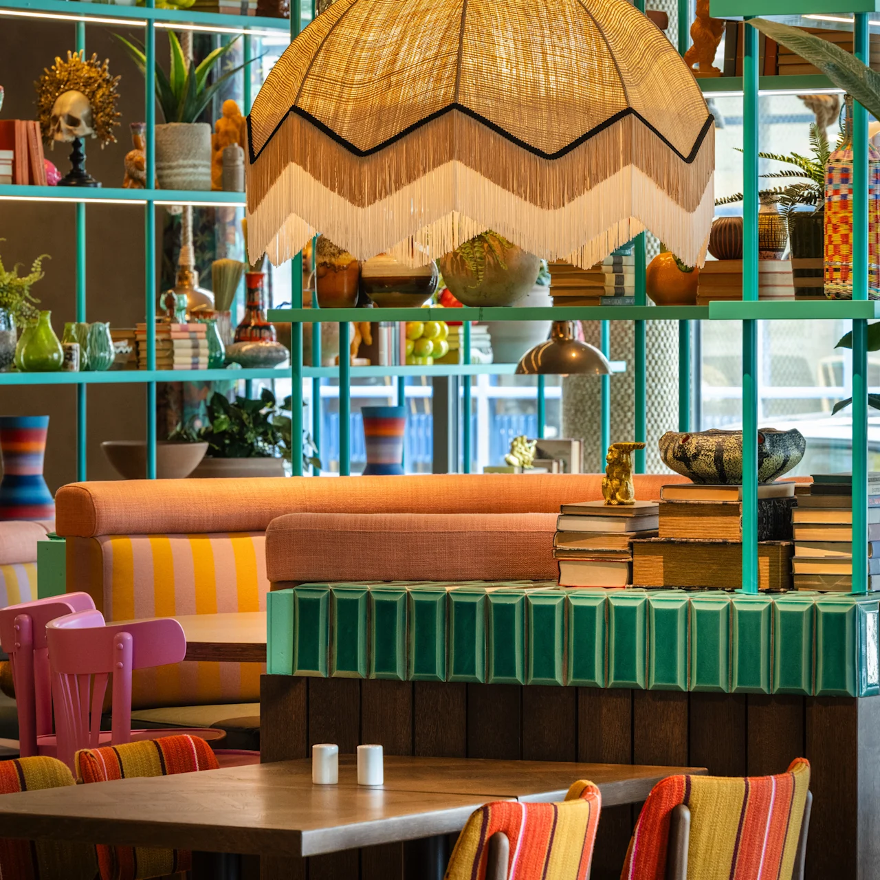Restaurant interior features colorful seating and tables. A large, decorative lamp hangs overhead. The area is surrounded by vibrant decor and plants, creating a lively and eclectic atmosphere.
