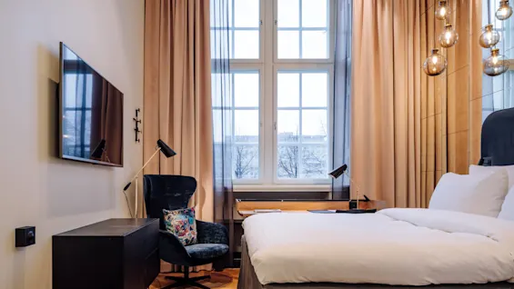 Television mounted on the wall, inactive, beside an armchair with a colorful pillow. Situated in a cozy bedroom featuring a large window and stylish light fixtures.