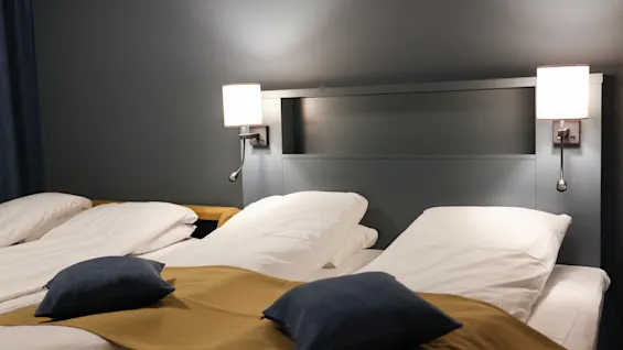 En prydligt bäddad dubbelsäng med vita lakan och kuddar, toppad med en brun sänglöpare och blå kuddar, flankeras av två väggmonterade lampor. Rummet har mörkblå väggar.