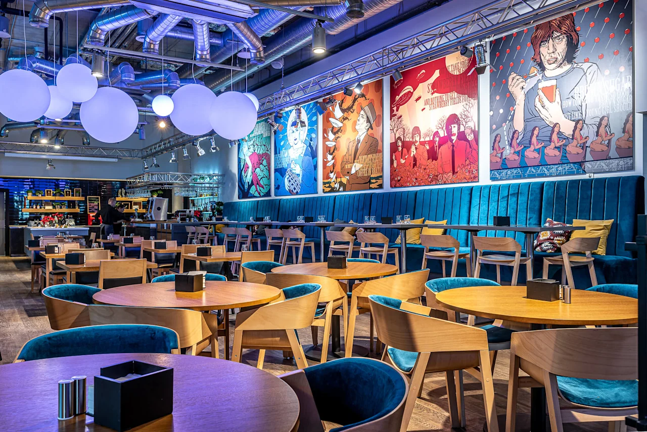 Stylish restaurant interior features circular wooden tables and turquoise chairs, beneath large round lights. Colorful pop art posters adorn the walls, creating a contemporary, vibrant atmosphere.