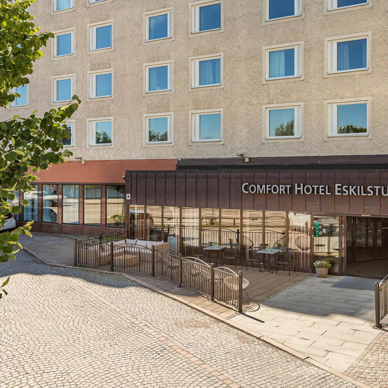 Comfort Hotel Eskilstunan sisäänkäynnissä on ulkoseating pöytineen ja tuoleineen, joka sijaitsee kadunkulmassa mukulakivipäällysteellä. Aluetta ympäröivät kaupunkirakennukset.