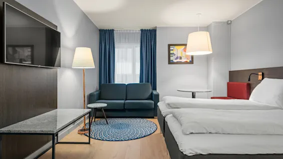 Et hotelværelse har en dobbeltseng, vægmonteret tv, sofa og et lille bord. Det har blå gardiner, en gulvlampe og moderne indretning.