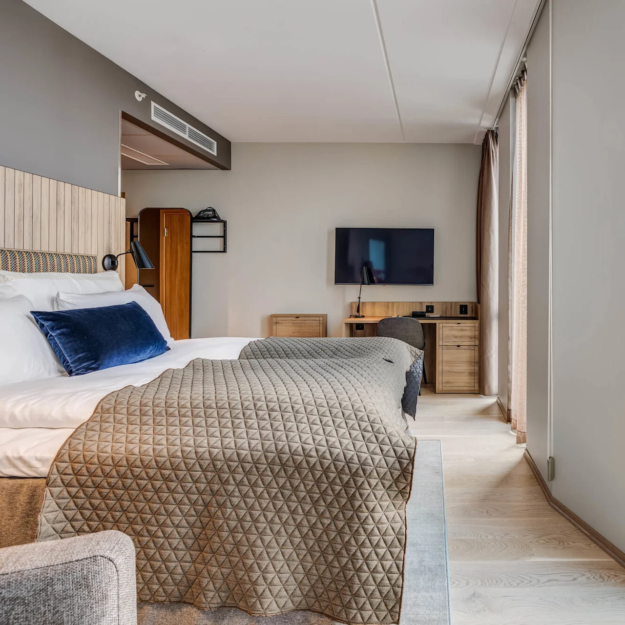 A neatly made bed with a gray quilt faces a wall-mounted TV. Large windows provide a panoramic city view, filling the modern room with natural light.