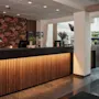 Reception skranke lyser varmt med vertikala trästänger; bakom en blommig vägg och datorer. Kontext inkluderar krukväxter, stora fönster, kafésittområde synligt i bakgrunden.