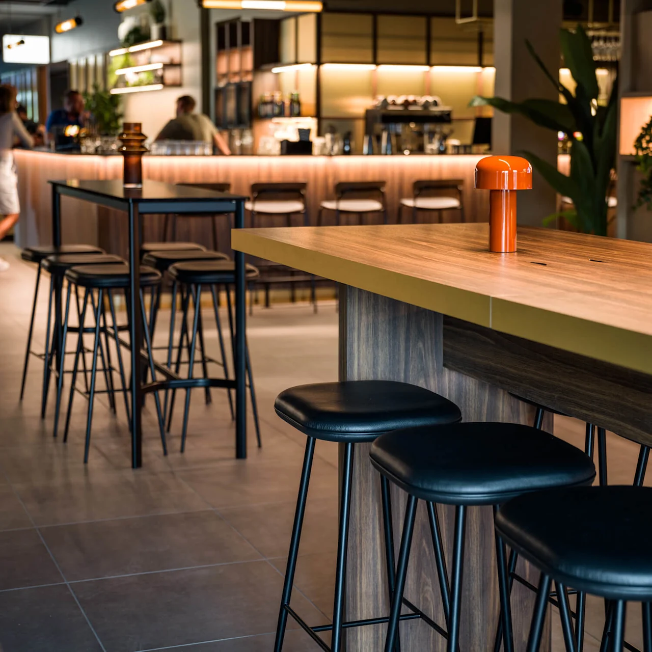 Bardisk med svarte krakker, med livlige oransje lamper på en treoverflate. Folk står og snakker i et varmt opplyst moderne bar-miljø.