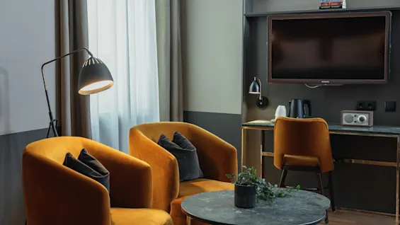 Et varmt og indbydende hotelværelse med komfortable siddepladser, et sofabord og et funktionelt skrivebordsområde med et fjernsyn.