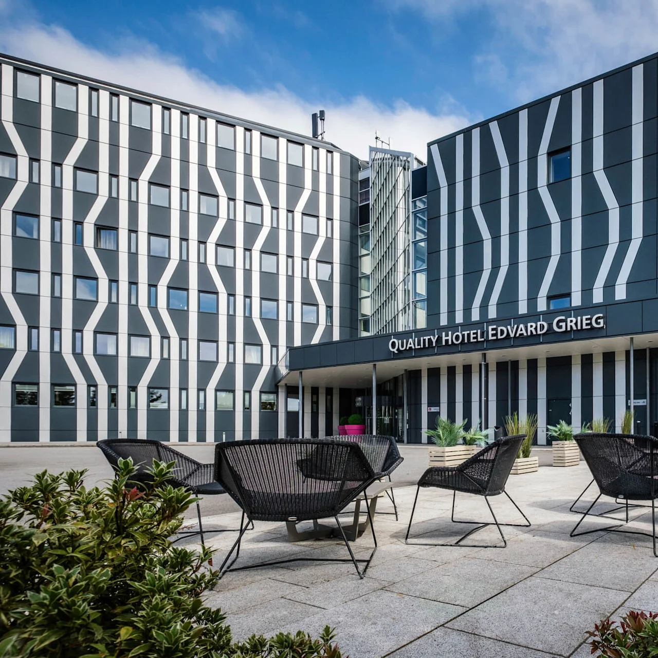 Moderne hotellbygning med stripet fasade, som viser skiltet "QUALITY HOTEL EDVARD GRIEG", omgitt av utemøbler og grønne planter under en delvis skyet himmel.