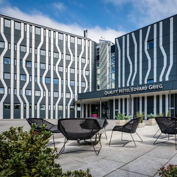 Moderne hotellbygning med stripet fasade, som viser skiltet "QUALITY HOTEL EDVARD GRIEG", omgitt av utemøbler og grønne planter under en delvis skyet himmel.
