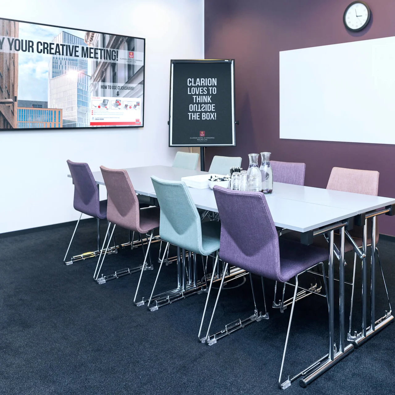Et konferanserom med et rektangulært bord omgitt av fargerike stoler. På veggen står det "ENJOY YOUR CREATIVE MEETING!" og "CLARION LOVES TO THINK OUTSIDE THE BOX!" En klokke og et skap er til stede.