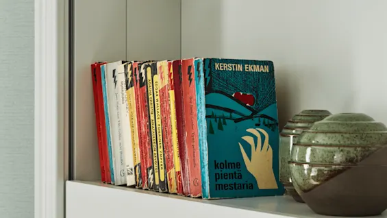 Bøker står oppreist på en hylle og viser fargerike rygger. En bok med tittelen “KERSTIN EKMAN kolme pientä mestaria” vender fremover. Keramiske potter står ved siden av.