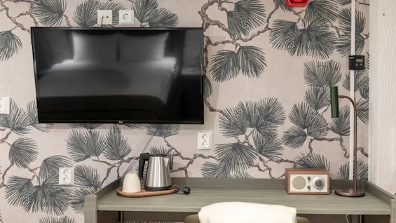 Moderni hotellihuone, jossa on seinälle asennettu televisio, virvokkeita ja viihdettä varten varustettu työpöytä sekä viihtyisä tuoli.