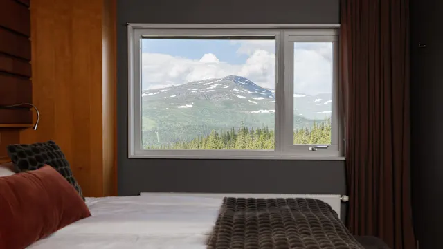 A bed with pillows faces a window showcasing a mountain view. The scene suggests a cozy interior with a nature-focused outlook.