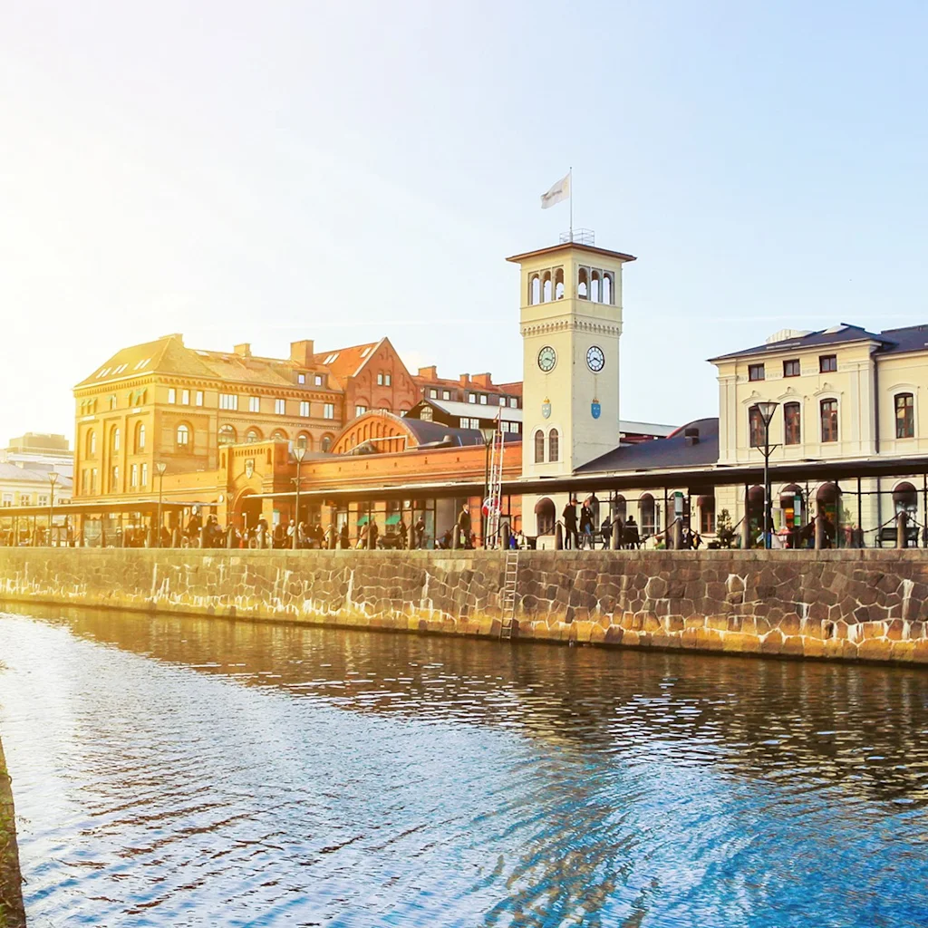 A canal flows alongside historic buildings with a clock tower, bathed in warm sunlight, reflecting a serene urban setting.