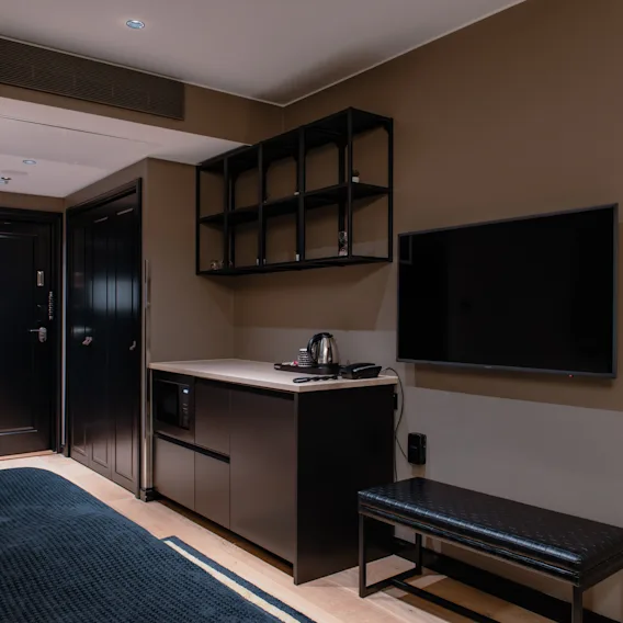 Hotellihuone, jossa viihtyisä sänky, kätevä minikeittiö mikroaaltouunilla ja vedenkeittimellä, iso TV ja tyylikäs penkki.