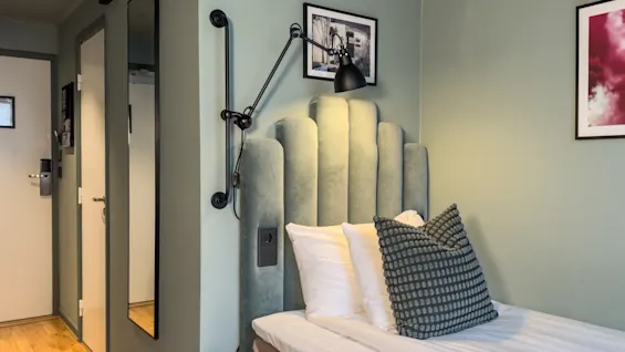 Et koselig hotellrom med en enkeltseng med en grønn polstret sengegavl, en stilig vegglampe og lysegrønne vegger.