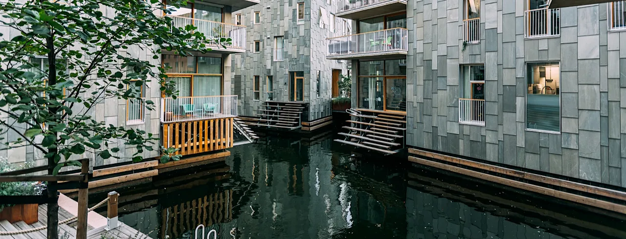 Moderne bygninger med balkoner med udsigt over en rolig kanal, med unikke flisebelagte facader og trapper, der fører ned til vandet.