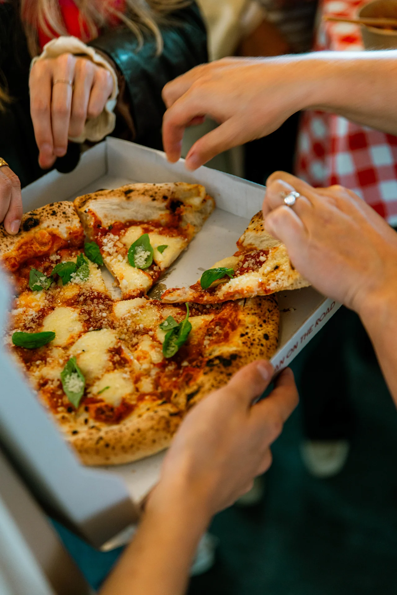En pizza med smält ost och basilika delas av flera händer från en låda, omgiven av människor i en avslappnad inomhusmiljö.