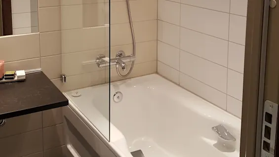 Badekar med veggmontert dusjhode i flislagt bad; glasskjerm separerer badekar fra servantområdet. To veggmonterte dispensere over badekar mot beige og hvite fliser.