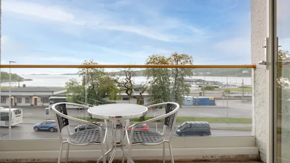 Et rundt hvidt bord med to metalstole står på en altan. Nedenfor, en vej med parkerede køretøjer; hinsides en udsigt til grønne områder, vand og en skyet himmel.