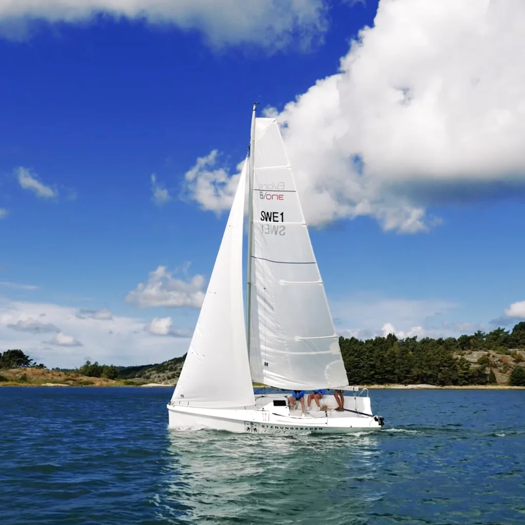 En vit segelbåt glider över lugnt blått vatten under en ljus himmel med fluffiga moln, med ett hus på en grön strand i fjärran.