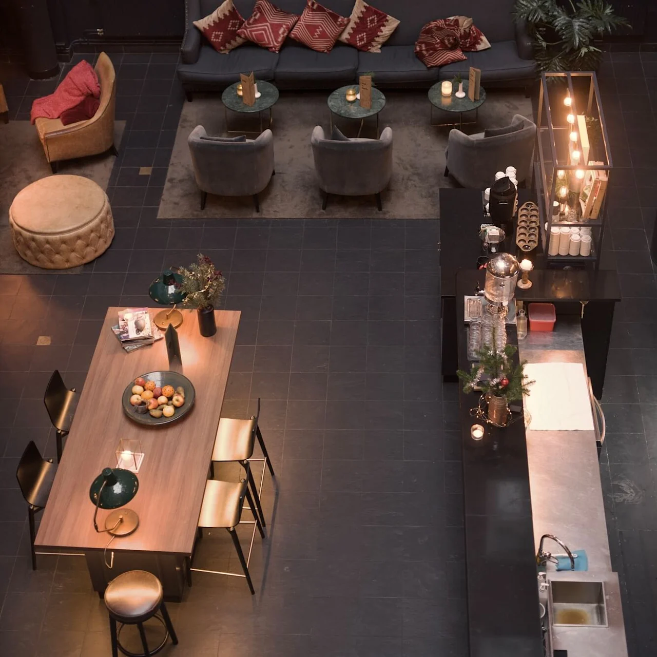 A cozy lounge area with sofas and armchairs, lit by warm lighting, in a modern indoor setting with black walls and floor tiles.