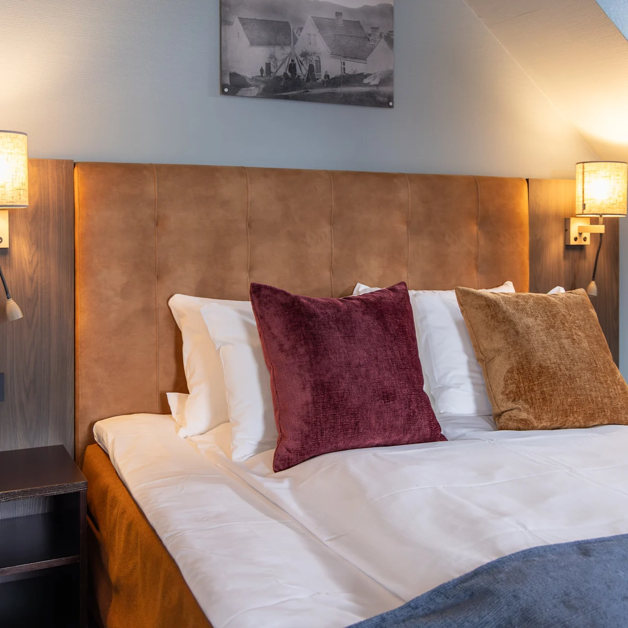 Bed with pillows sits in a cozy room. Nightstands with lamps flank the bed, casting warm light over a headboard. A monochrome picture hangs above, adding decoration.