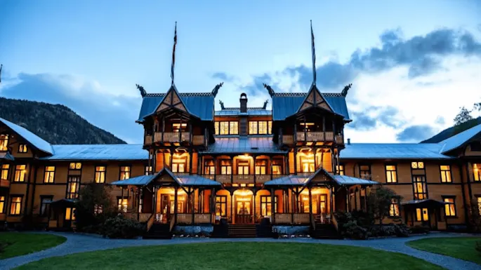 An ornate, illuminated wooden building with towers stands against a twilight sky, set amid grassy lawns and mountains in the background.