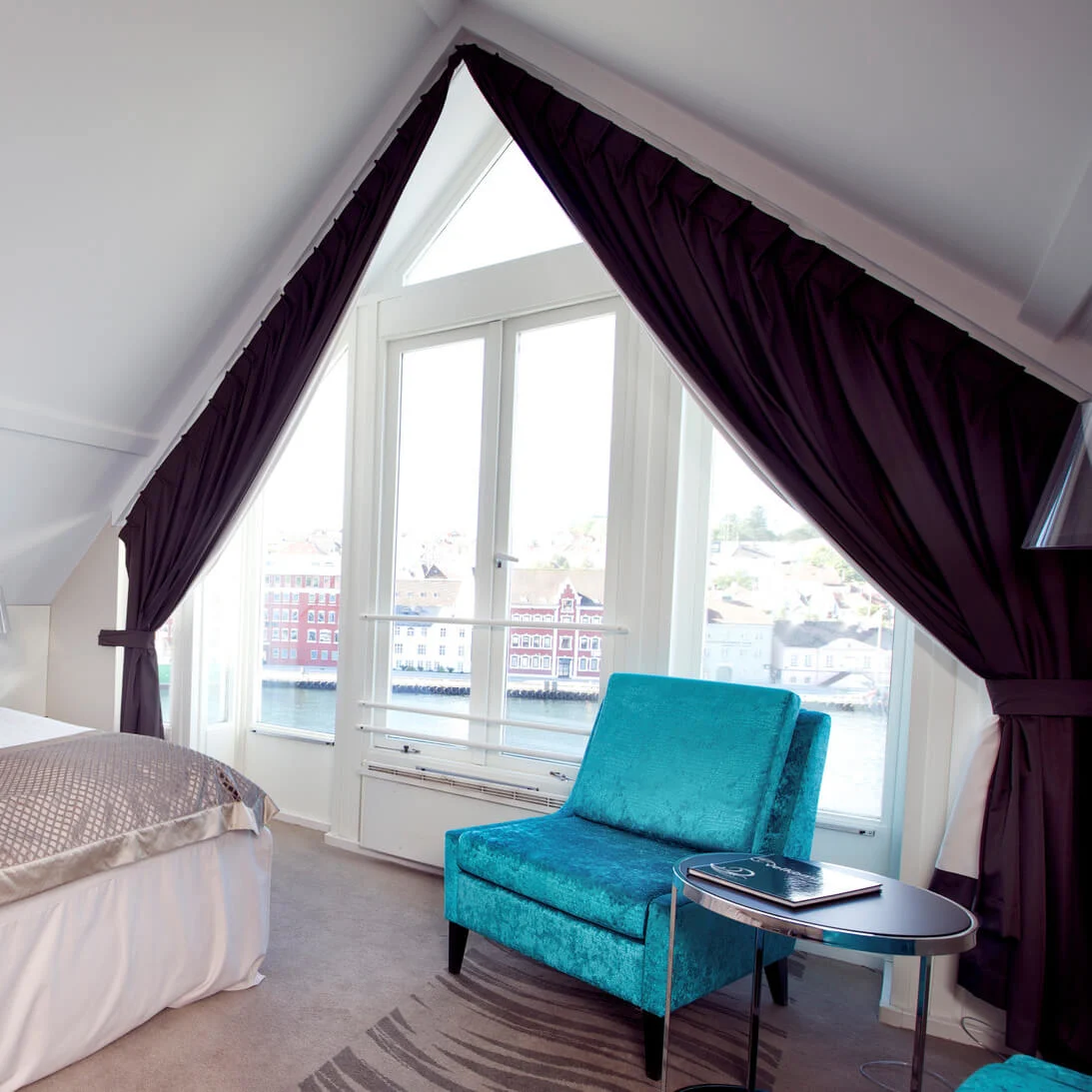 A well-lit attic bedroom with an inclined ceiling, large windows overlooking a body of water and buildings, plush bedding, and elegant furniture.
