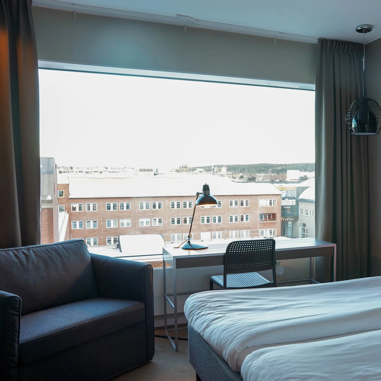 En seng med et orange sengegærde; et skrivebord ved siden af, med en lampe, vender mod et stort vindue med udsigt over en by med murstensbygninger.