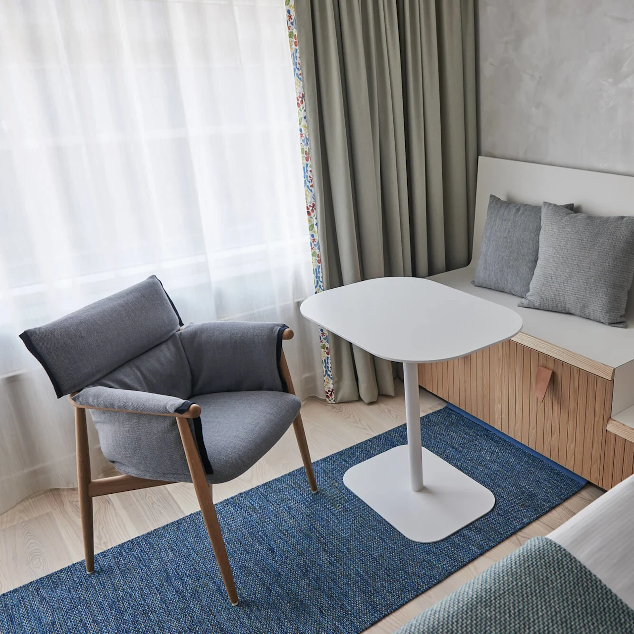 A modern room features a gray armchair beside a white table, with a bed and built-in seating area. Floor-length curtains frame a window, complementing the minimalist decor.