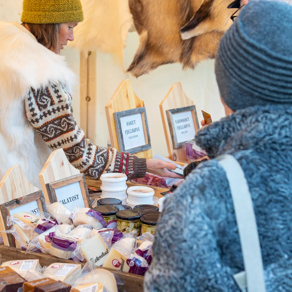 Two people at a market stall, one serving and one browsing food products. 