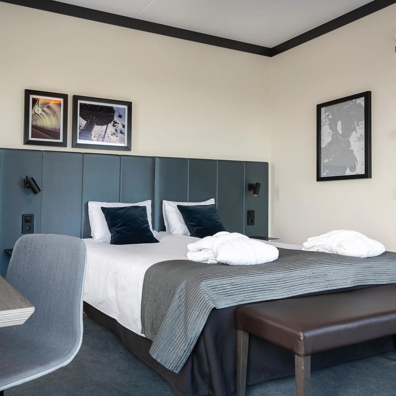 A neatly arranged hotel room features a double bed with white linens and dark cushions, under framed pictures on a green headboard. Nearby, a desk holds a coffee machine.
