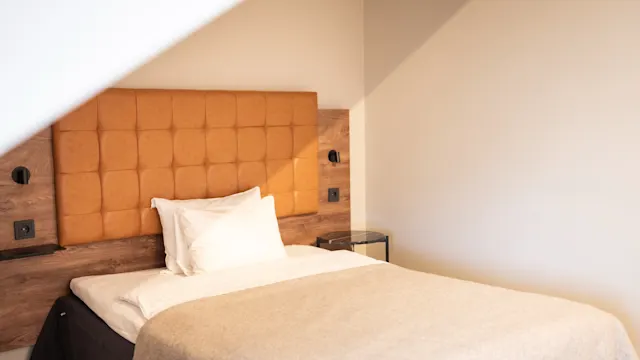 En seng med et beige tæppe og hvide puder står mod en væg, der har en polstret sengegavl. Blødt lys kommer ind i rummet og forbedrer den hyggelige atmosfære.