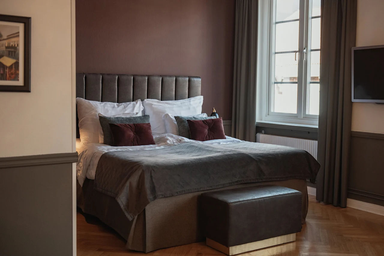 Ett mysigt hotellrum med en stor säng, varma bruna väggar och ett ljust fönster. En bekväm bänk står vid fotändan av sängen.