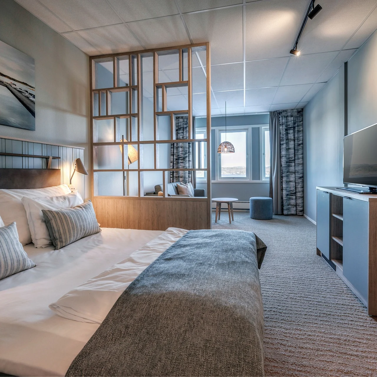A bed with white linens and patterned pillows stands against a partition. A TV is on a cabinet opposite, with a sitting area near large windows.