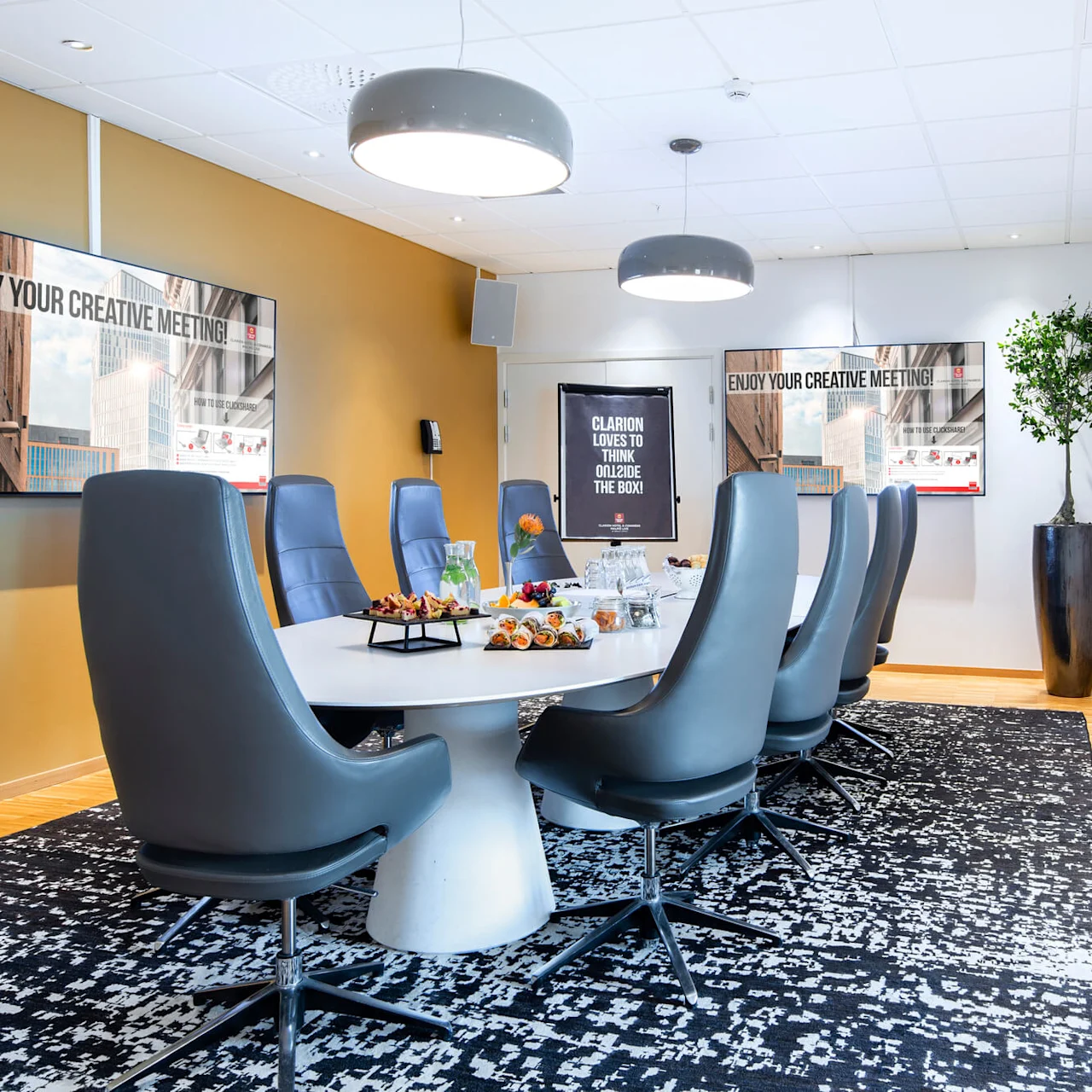Et moderne konferanserom med et stort ovalt bord omgitt av grå stoler. Bordet har snacks og drikke. Veggskjermer viser "ENJOY YOUR CREATIVE MEETING!". En potteplante står i nærheten.