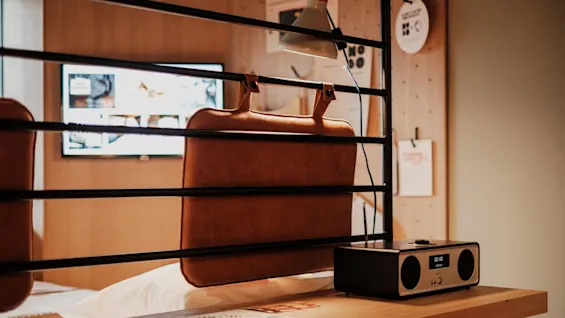 A leather backrest hangs from a black bed frame, with a table underneath. A digital radio reads "00:00." A wall-mounted TV and various papers are in the background.